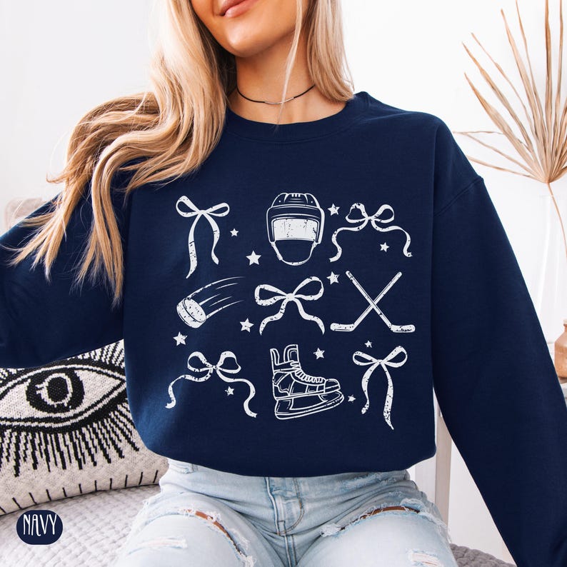 May include: A navy blue sweatshirt with a white graphic design featuring hockey-themed illustrations, including a hockey helmet, skates, sticks, and bows.
