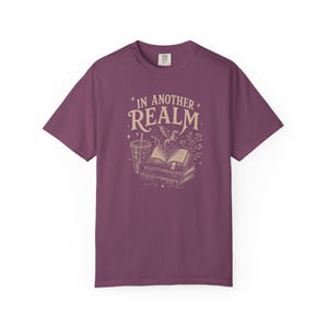 May include: A plum-colored short-sleeve t-shirt featuring a cream-colored graphic print. The design displays a stack of books, a drink cup, and a small dragon with the text IN ANOTHER REALM surrounded by stars and floral elements.