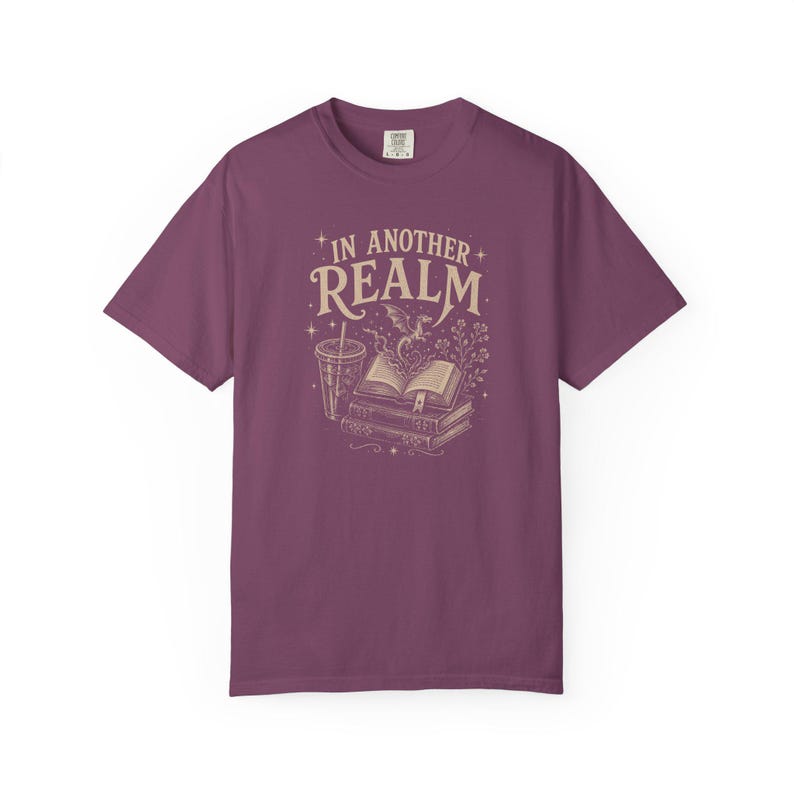 May include: A plum-colored short-sleeve t-shirt featuring a cream-colored graphic print. The design displays a stack of books, a drink cup, and a small dragon with the text IN ANOTHER REALM surrounded by stars and floral elements.