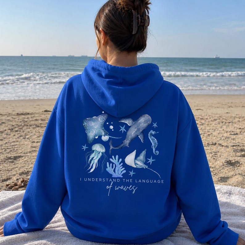 Pu&ograve; includere: Felpa con cappuccio blu reale con grafica di vita marina. Il design include una razza, uno squalo, un cavalluccio marino, una medusa e coralli, con la scritta "I understand the language of waves". La felpa &egrave; realizzata in materiale morbido.