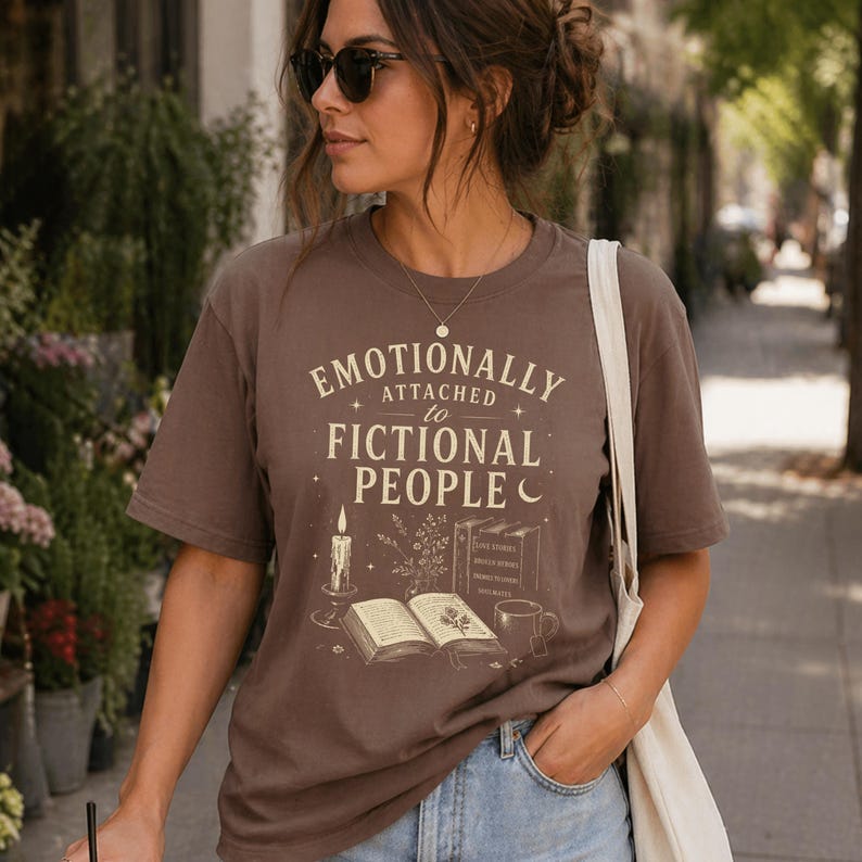May include: A brown t-shirt featuring the phrase "Emotionally Attached to Fictional People." The design includes a candle, open book, coffee cup, and books. The shirt is worn by a person wearing sunglasses and a necklace.