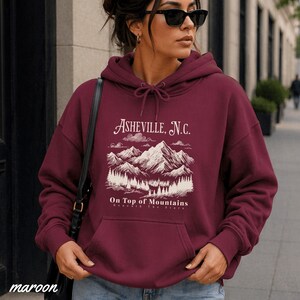 May include: Maroon hoodie with a front pocket and drawstring. The white graphic on the front reads "Asheville, N.C." above a mountain scene and the words "On Top of Mountains Beneath The Stars". The word "maroon" is printed at the bottom.