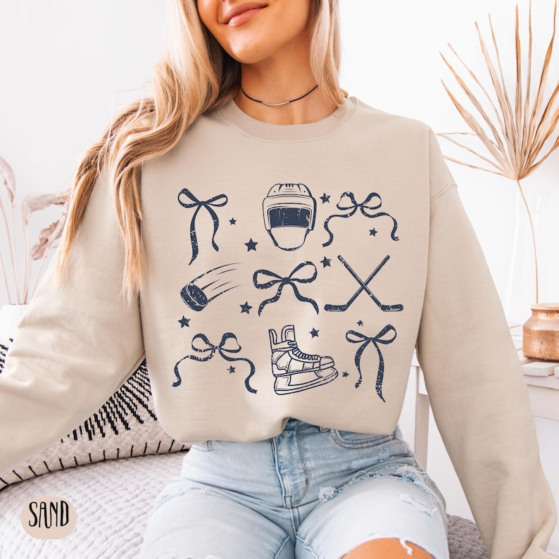 May include: A beige sweatshirt with a hockey-themed design. The design features a hockey helmet, hockey sticks, a puck, a pair of ice skates, and bows. The design is in a dark blue color.