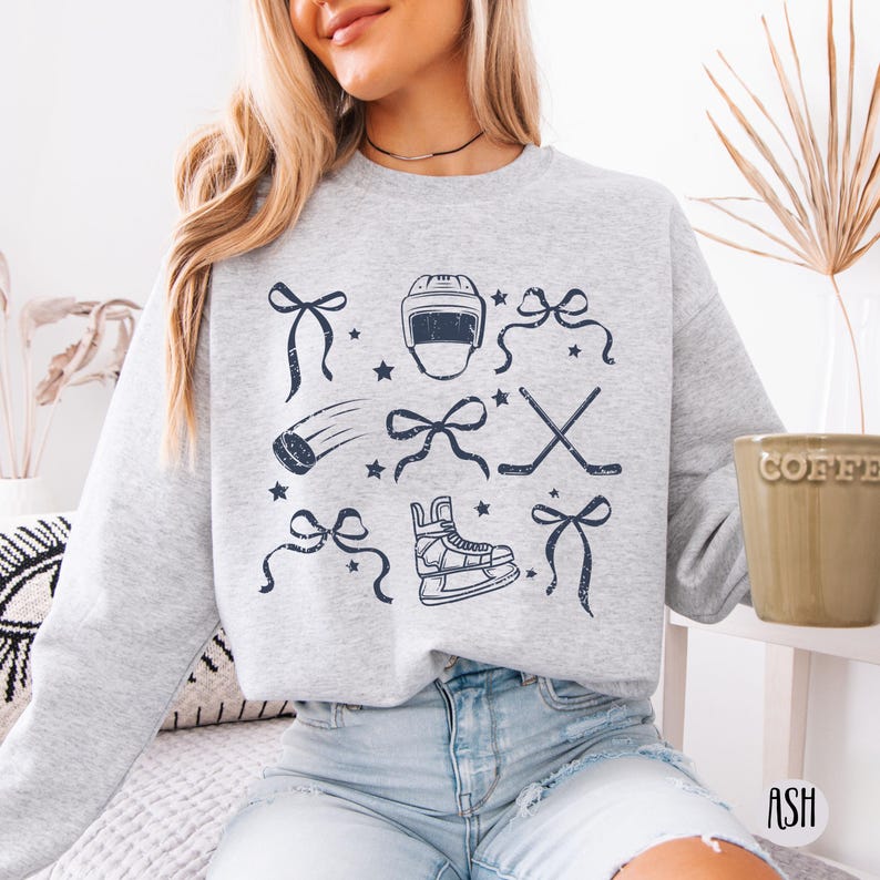 May include: A gray sweatshirt with a blue graphic print of hockey-themed items, including a hockey helmet, skates, sticks, and bows. The sweatshirt is worn by a person sitting down.