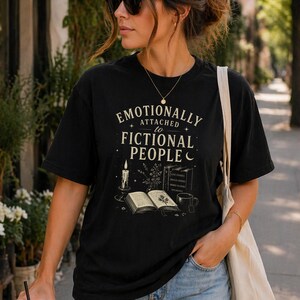 May include: Black t-shirt featuring the phrase "Emotionally Attached to Fictional People". The design includes a candle, an open book, and a mug. The shirt is worn by a person wearing sunglasses and a necklace.