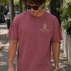 May include: A dusty rose short-sleeve t-shirt featuring a small graphic on the upper left chest. The graphic depicts a vintage lantern above the text "TRUST THE TRAIL" and "ISAIAH 41:10" printed in a light, neutral color on the fabric.