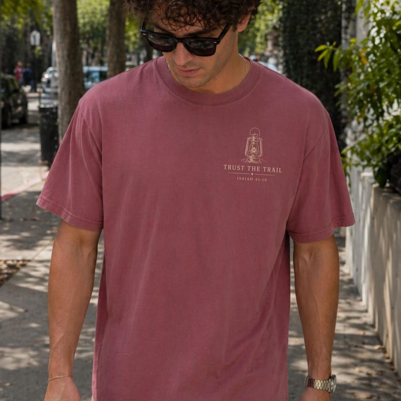 May include: A dusty rose short-sleeve t-shirt featuring a small graphic on the upper left chest. The graphic depicts a vintage lantern above the text "TRUST THE TRAIL" and "ISAIAH 41:10" printed in a light, neutral color on the fabric.