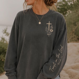 May include: A charcoal gray long-sleeve crewneck sweatshirt featuring a beige graphic on the upper left chest of a cross surrounded by flowers. The left sleeve displays the beige text Trust in the Lord and the reference Proverbs 3:5.