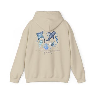 Pu&ograve; includere: Felpa con cappuccio beige con un disegno ad acquerello di vita marina, tra cui una manta, una medusa e un cavalluccio marino. Il testo "I understand the language of waves" &egrave; stampato sotto. La felpa ha un cappuccio con coulisse e maniche lunghe.