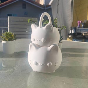 Peut inclure: Un récipient blanc en forme de chat avec une poignée, empilé sur un autre récipient similaire. Les récipients ont des traits de visage et sont posés sur une table en verre. De petites plantes en pot sont à l'arrière-plan.