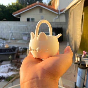 May include: An off-white cat-shaped planter with a handle. The planter features rounded ears, a nose, and whiskers. The planter is held in a hand, with a blurred outdoor background visible.