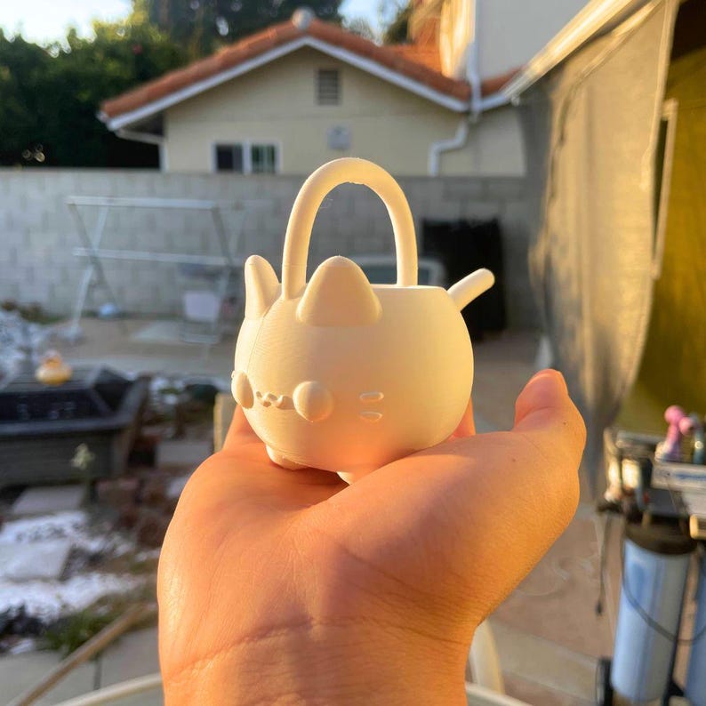May include: An off-white cat-shaped planter with a handle. The planter features rounded ears, a nose, and whiskers. The planter is held in a hand, with a blurred outdoor background visible.