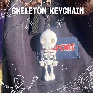 Pode incluir: Chaveiro de esqueleto branco com cabeça em forma de caveira e membros articulados. O chaveiro está pendurado em uma mochila preta. As palavras "SKELETON KEYCHAIN" estão escritas em branco acima do chaveiro. A mochila tem um patch azul que diz "SPORT".