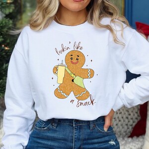 May include: White sweatshirt with a gingerbread man graphic holding a drink and a bag. The text "lookin' like a snack" is written above and below the gingerbread man. The sweatshirt is a casual style.