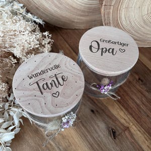 Könnte beinhalten: Zwei Gläser mit Holzdeckeln. Ein Deckel trägt die Aufschrift "Wundervolle Tante" mit einem Herz, der andere "Großartiger Opa" mit einem Herz. Jedes Glas ist mit Bindfaden und kleinen Blumengestecken umwickelt.