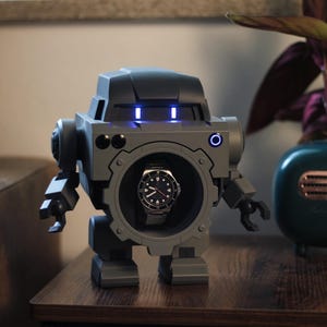 May include: A gray robot-shaped watch winder with a watch displayed in the center. The robot has blue lights on its head and mechanical arms and legs. A teal planter with a plant is to the right.