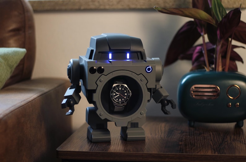 May include: A gray robot-shaped watch winder with a watch displayed in the center. The robot has blue lights on its head and mechanical arms and legs. A teal planter with a plant is to the right.