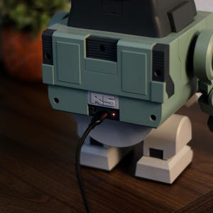 May include: A retro-style robot-shaped device in shades of green, gray, and black, with a charging cable plugged in. The robot has a boxy body, articulated arm, and a small red light indicating power. The device is on a wooden surface.