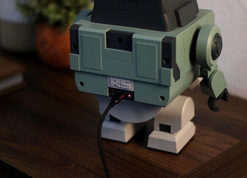 May include: A retro-style robot-shaped device in shades of green, gray, and black, with a charging cable plugged in. The robot has a boxy body, articulated arm, and a small red light indicating power. The device is on a wooden surface.