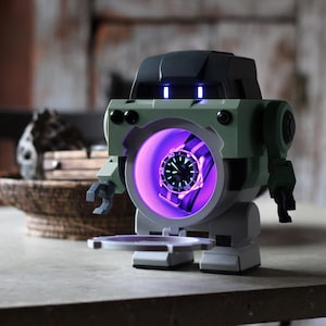 May include: A robot-shaped watch display case in shades of green, gray, and black. The case has a circular opening with a watch illuminated by a purple light. The robot has a tray extending from its base.