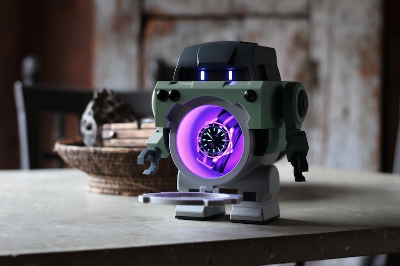 May include: A robot-shaped watch display case in shades of green, gray, and black. The case has a circular opening with a watch illuminated by a purple light. The robot has a tray extending from its base.