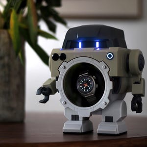 Pode incluir: Um expositor de relógios em forma de robô bege e cinza, com um relógio visível no centro. O robô tem luzes azuis e uma abertura circular. O relógio tem um mostrador preto e uma pulseira prateada. O robô está sobre uma superfície de madeira.