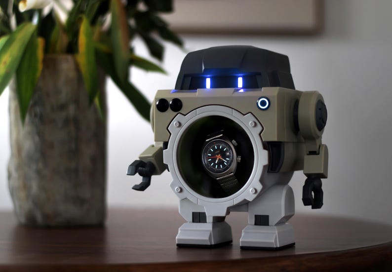 May include: A robot-shaped watch display case in beige and gray, with a watch visible in the center. The robot has blue lights and a circular opening. The watch has a black face and a silver band. The robot is on a wooden surface.