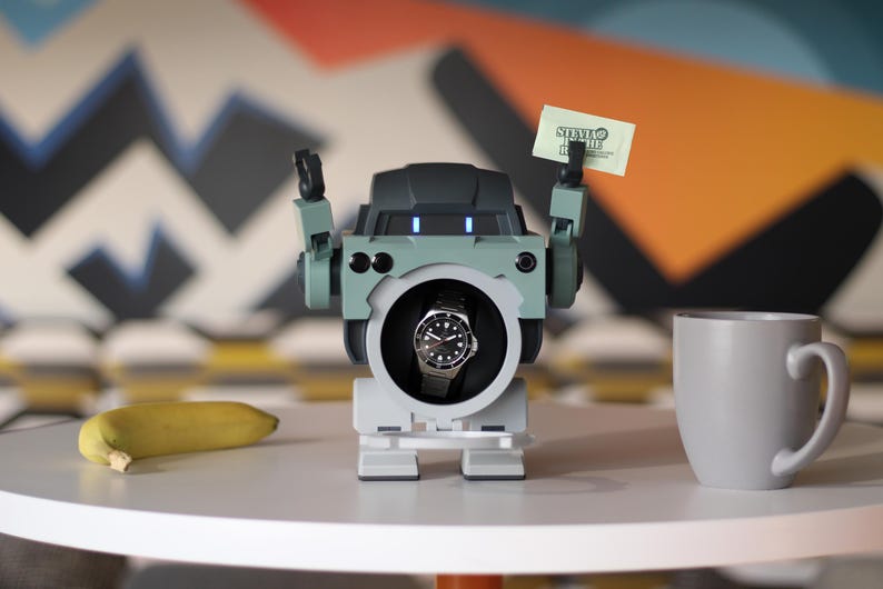 May include: A robot-shaped watch winder with a black and silver watch inside. The robot has green and gray accents and holds a small card. A banana and a gray mug sit on the table.