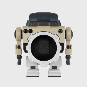 May include: A beige and black robot-shaped watch winder. The winder has a large circular opening in the center, with a white gear-like frame. The robot has tan arms and legs, and a black head.