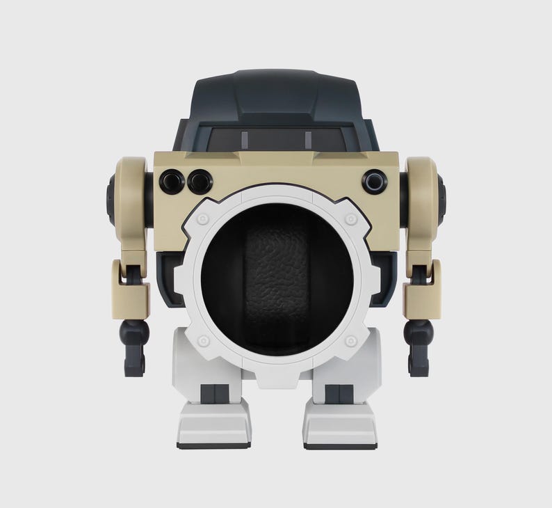 May include: A beige and black robot-shaped watch winder. The winder has a large circular opening in the center, with a white gear-like frame. The robot has tan arms and legs, and a black head.