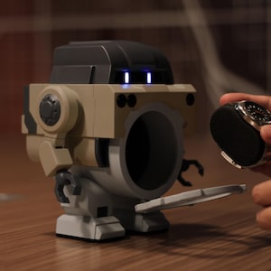 May include: A tan and black robot-shaped speaker with a large circular opening. The robot has a black top and blue lights. A person is holding a black watch with a silver bezel near the speaker.