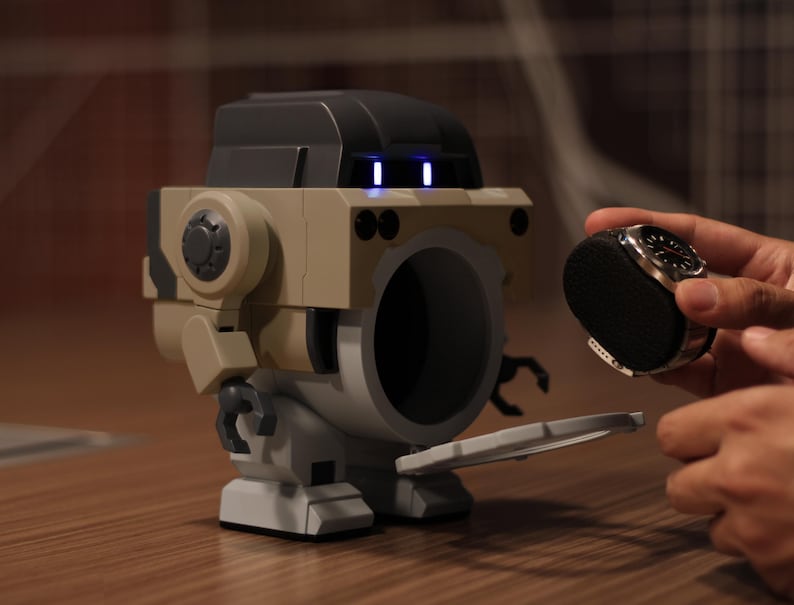 May include: A tan and black robot-shaped speaker with a large circular opening. The robot has a black top and blue lights. A person is holding a black watch with a silver bezel near the speaker.