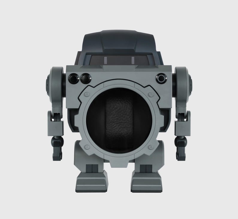 May include: A gray and black robot-shaped watch winder. The winder has a large circular opening in the center, with a dark interior. It features articulated arms and legs, and a dark gray top section with a window-like design.