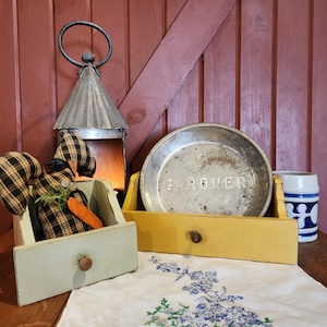 May include: Two wooden drawers with a plaid fabric bunny inside the left drawer. The right drawer has a metal pie plate with the word "GARDENER" printed on it. A blue and white ceramic mug is on the right side of the image.