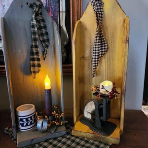 May include: Two wooden candle holders with a rustic, primitive style. The holder on the left is painted blue and features a candle, a small ceramic mug, and a metal ring. The holder on the right is painted yellow and features a small scale with a red berry garland.