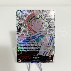 May include: A holographic trading card featuring Koby from the One Piece anime. The card has a colorful design with a manga-style illustration of Koby, text, and a holographic background. The card is displayed on a clear stand.
