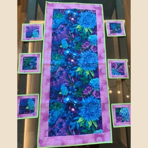 Puede incluir: Un camino de mesa morado y verde con un patrón floral con flores azules. El camino de mesa está rodeado de seis posavasos a juego.