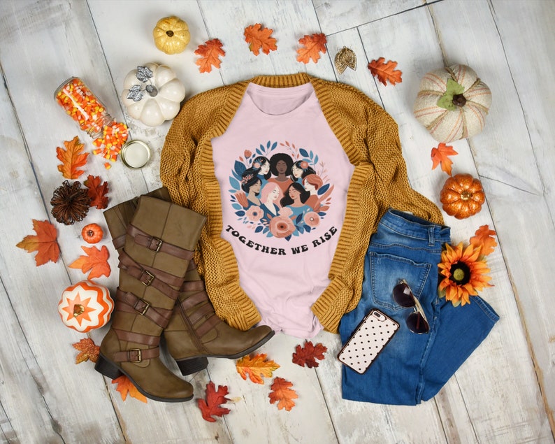 K&ouml;nnte beinhalten: Rosa T-Shirt mit einem Motiv eines Blumenkranzes mit verschiedenen Frauen darin. Der Text "Together We Rise" befindet sich unter dem Kranz. Das Shirt ist mit einem braunen Cardigan, einer blauen Jeans, braunen Stiefeln und Herbstdeko gestylt.