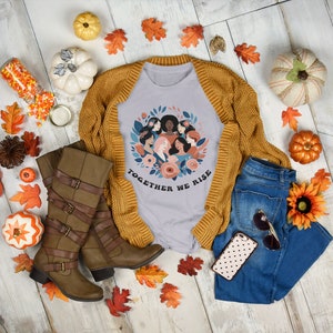 K&ouml;nnte beinhalten: Ein hellgraues T-Shirt mit einem Grafikdruck eines Kreises von Frauen mit Blumen und dem Text "Together We Rise". Das Shirt wird mit einem braunen Cardigan, braunen Stiefeln, einer blauen Jeans, einer Handyh&uuml;lle mit Punkten und Herbstdeko kombiniert.