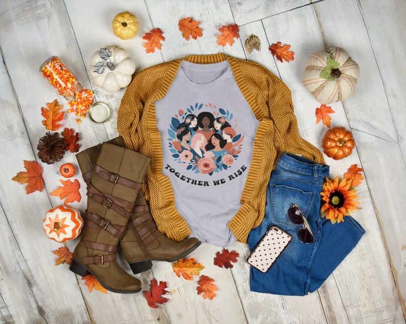 K&ouml;nnte beinhalten: Ein hellgraues T-Shirt mit einem Grafikdruck eines Kreises von Frauen mit Blumen und dem Text "Together We Rise". Das Shirt wird mit einem braunen Cardigan, braunen Stiefeln, einer blauen Jeans, einer Handyh&uuml;lle mit Punkten und Herbstdeko kombiniert.