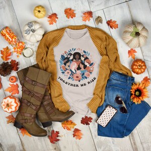 K&ouml;nnte beinhalten: Ein beiges T-Shirt mit einem Grafikdruck eines Kreises von Frauen mit Blumen und dem Text "Together We Rise". Das Shirt ist mit einem braunen Cardigan, einer blauen Jeans, braunen Stiefeln und Herbstdeko gestylt.