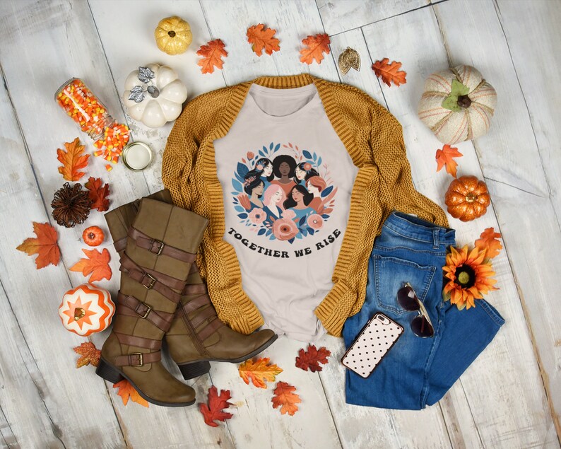 K&ouml;nnte beinhalten: Ein beiges T-Shirt mit einem Grafikdruck eines Kreises von Frauen mit Blumen und dem Text "Together We Rise". Das Shirt ist mit einem braunen Cardigan, einer blauen Jeans, braunen Stiefeln und Herbstdeko gestylt.