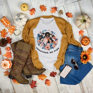 K&ouml;nnte beinhalten: Wei&szlig;es T-Shirt mit einem Grafikdruck eines Kreises von Frauen mit Blumen und dem Text "Together We Rise". Das Shirt wird auf einer Holzoberfl&auml;che mit Herbstdekorationen wie K&uuml;rbissen, Bl&auml;ttern und einer Sonnenblume pr&auml;sentiert.