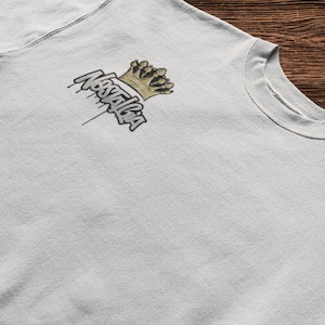 Puede incluir: Una camiseta gris claro con un gráfico negro y dorado de una corona y la palabra "Nostalgia" goteando de la corona.