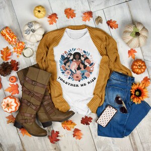 K&ouml;nnte beinhalten: Wei&szlig;es T-Shirt mit einem Grafikdruck einer Frauenkr&auml;nze mit dem Text "Together We Rise". Das Shirt wird auf einem wei&szlig;en Holzgrund mit Herbstdeko, darunter K&uuml;rbisse, Bl&auml;tter und eine braune Strickjacke, pr&auml;sentiert.
