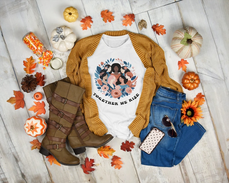 K&ouml;nnte beinhalten: Wei&szlig;es T-Shirt mit einem Grafikdruck einer Frauenkr&auml;nze mit dem Text "Together We Rise". Das Shirt wird auf einem wei&szlig;en Holzgrund mit Herbstdeko, darunter K&uuml;rbisse, Bl&auml;tter und eine braune Strickjacke, pr&auml;sentiert.