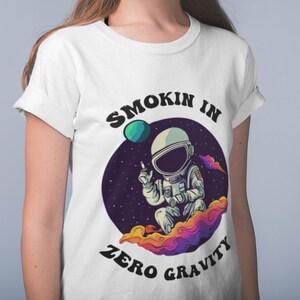 Peut inclure: T-shirt blanc avec un graphique d'un astronaute fumant dans l'espace. Le texte "Smokin in Zero Gravity" est imprimé sur le t-shirt.