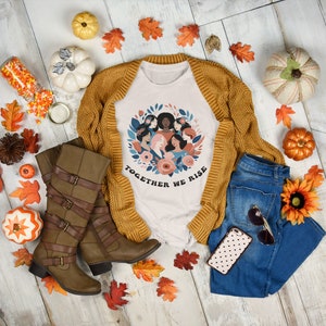K&ouml;nnte beinhalten: Ein wei&szlig;es T-Shirt mit einem Motiv eines Kranzes aus verschiedenen Frauen mit dem Text "Together We Rise". Das Shirt wird mit einem braunen Cardigan, einer blauen Jeans, braunen Stiefeln und Herbstdeko kombiniert.
