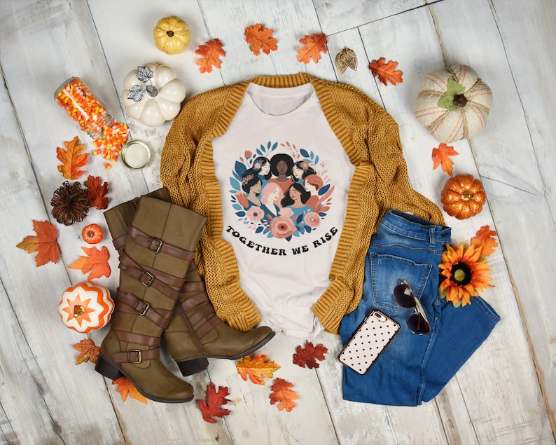 K&ouml;nnte beinhalten: Ein wei&szlig;es T-Shirt mit einem Motiv eines Kranzes aus verschiedenen Frauen mit dem Text "Together We Rise". Das Shirt wird mit einem braunen Cardigan, einer blauen Jeans, braunen Stiefeln und Herbstdeko kombiniert.