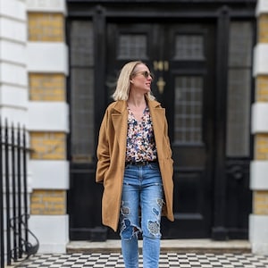 Woman coming out of a house in London wearing her floral wrap blouse with access for breastfeeding or pumping. Styled with Jeans and a camel coloured coat and glasses.